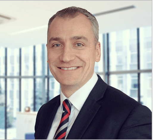 Andy McAllister smiles broadly, in a navy suit and red and grey striped tie