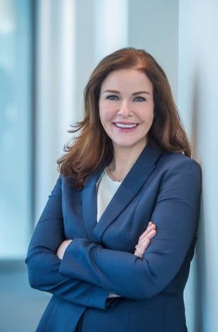 Angela Noon smiling, leans against a wall with arms folded