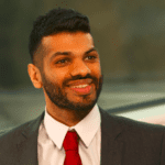 Deven Vedhera smiling in a grey suit and red tie, looking to the side of the camera