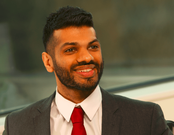 Deven Vedhera smiling in a grey suit and red tie, looking to the side of the camera