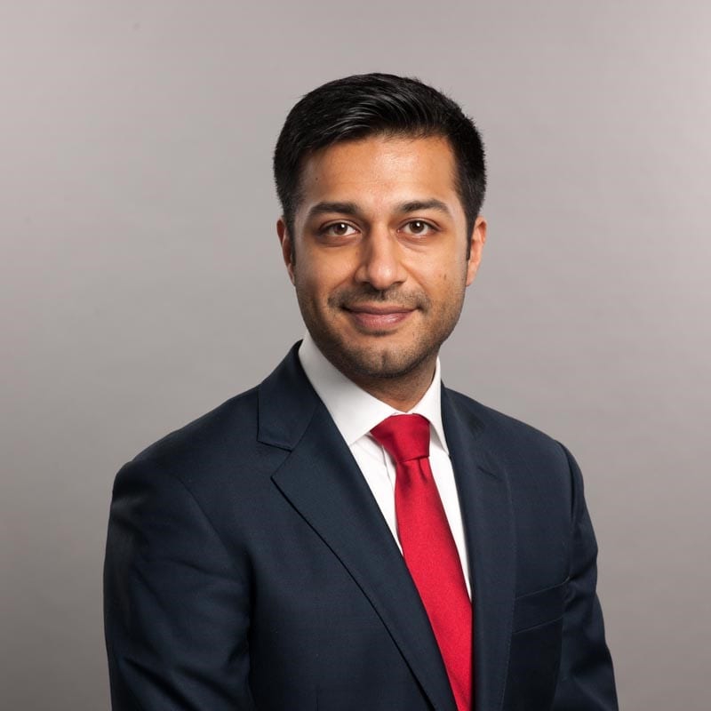 Manroop Khela in a dark suit and red tie looks at the camera